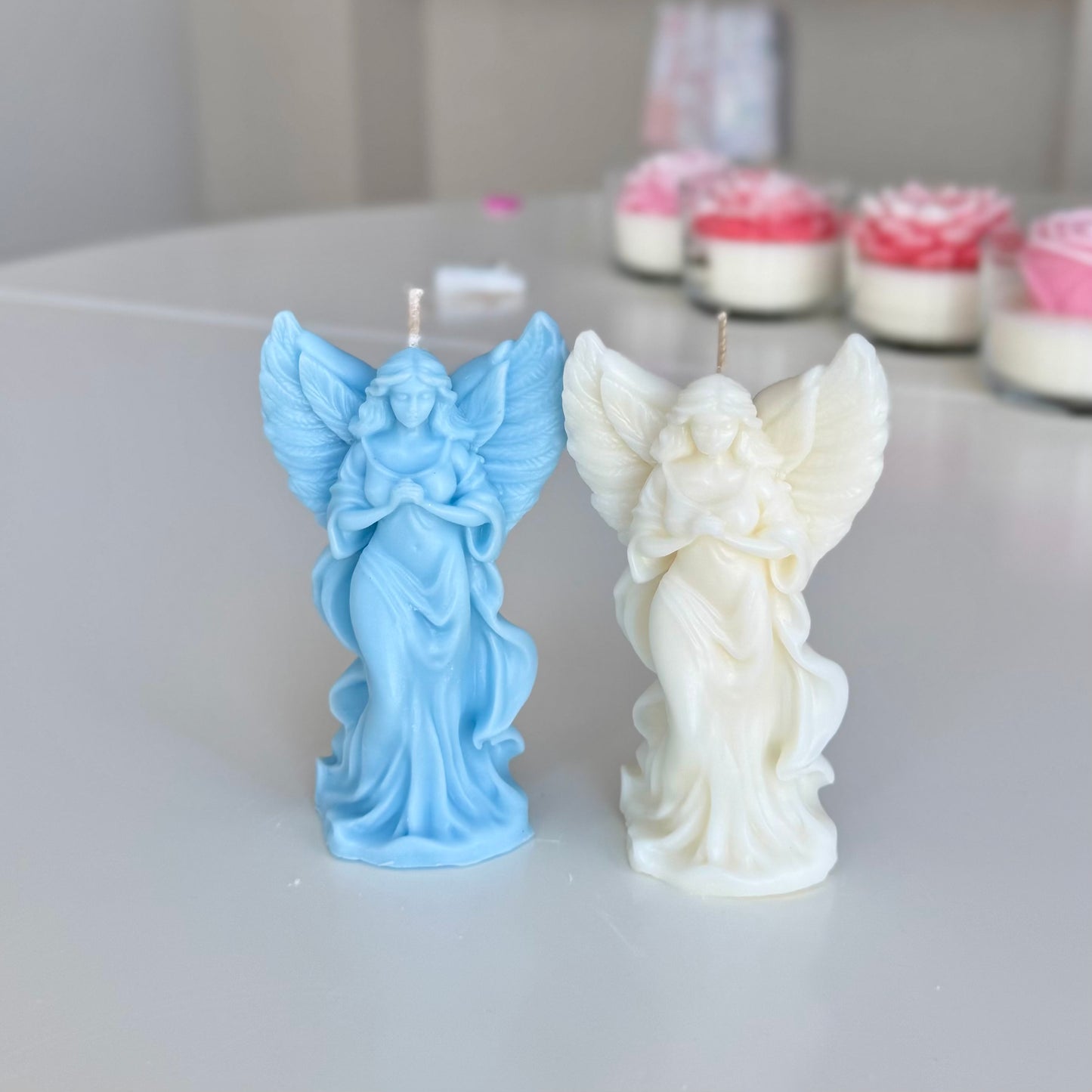 Two angel-shaped candles, one blue and one white, on a light surface with blurred background.

