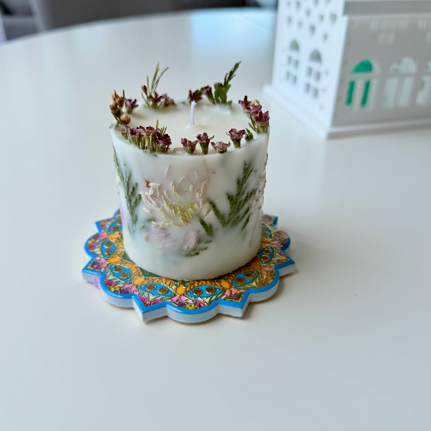 Decorative candle with floral elements on a colorful coaster, placed on a white surface.
