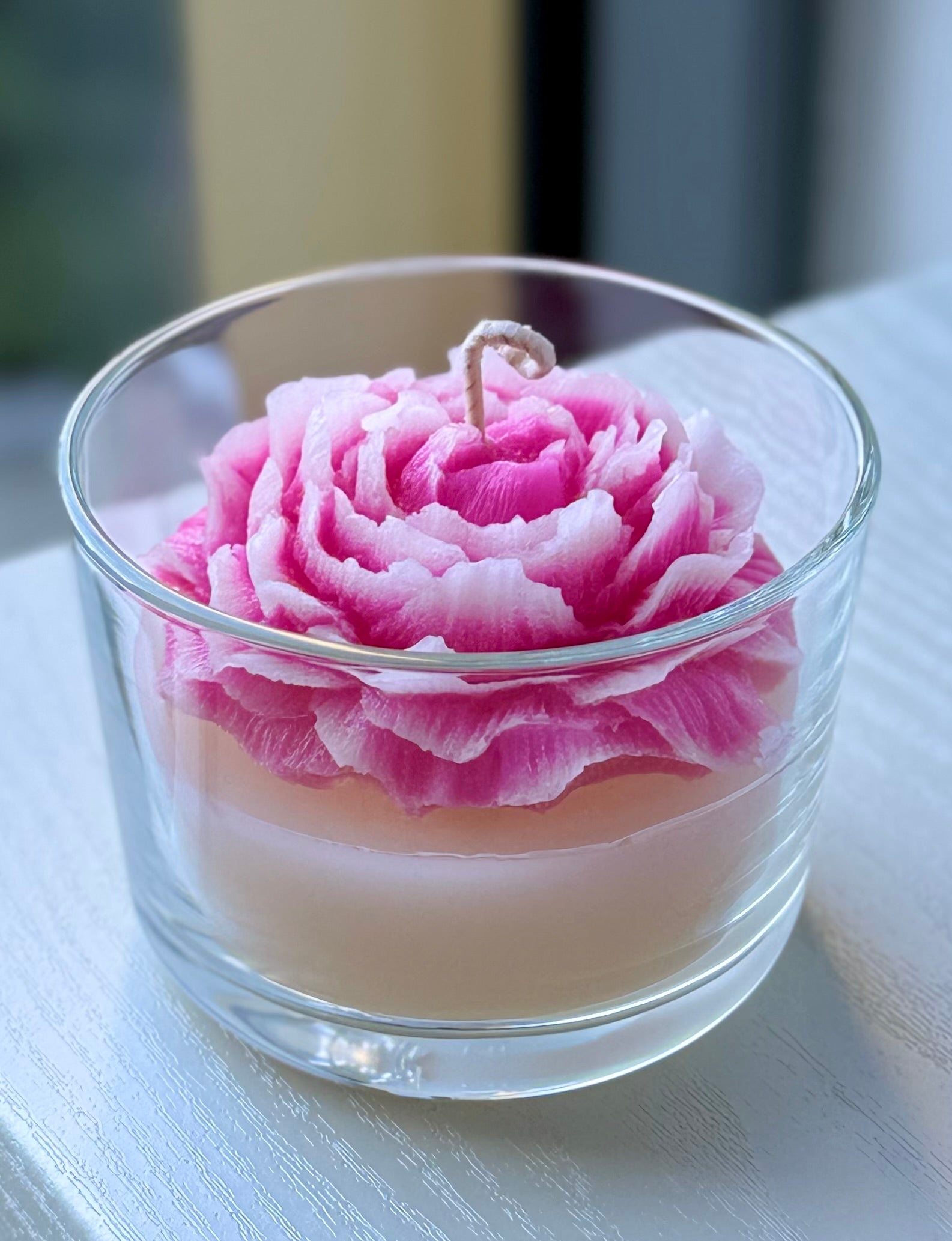 Handmade peony carved candle in pink and white flower design set in a clear glass cup on a light surface with blurred background.