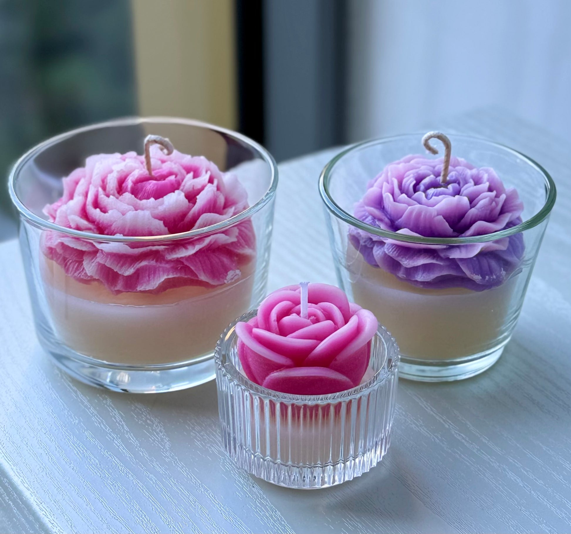 Set of three handmade peony carved candles in pink and purple colours with a small candle in front, displayed in glass holders on a white surface with blurred background.